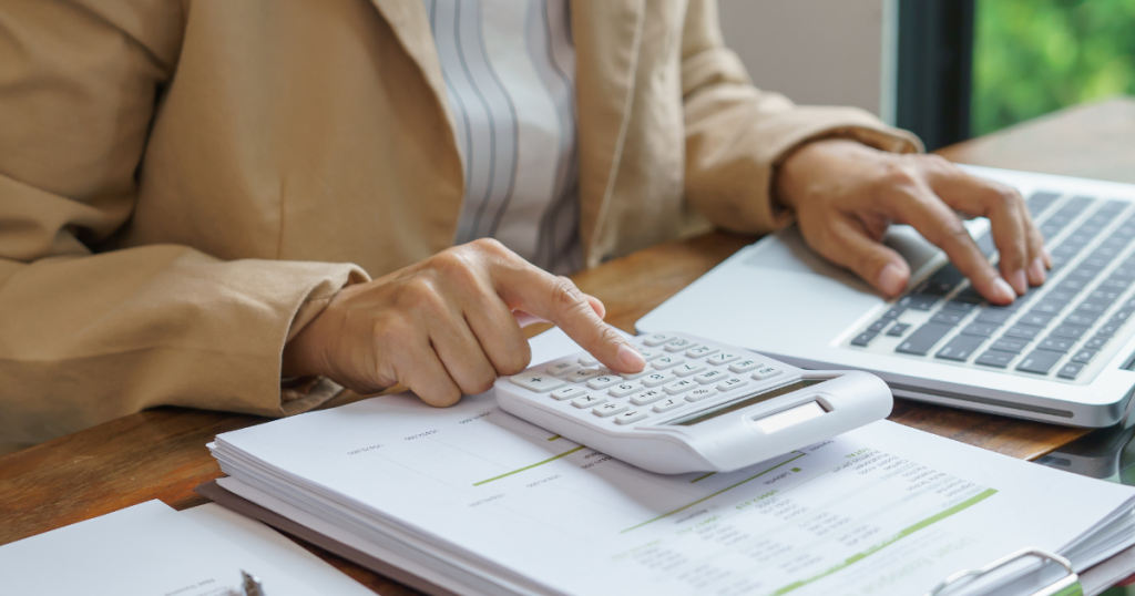A person calculating finances with a calculator and laptop on a desk indoors.